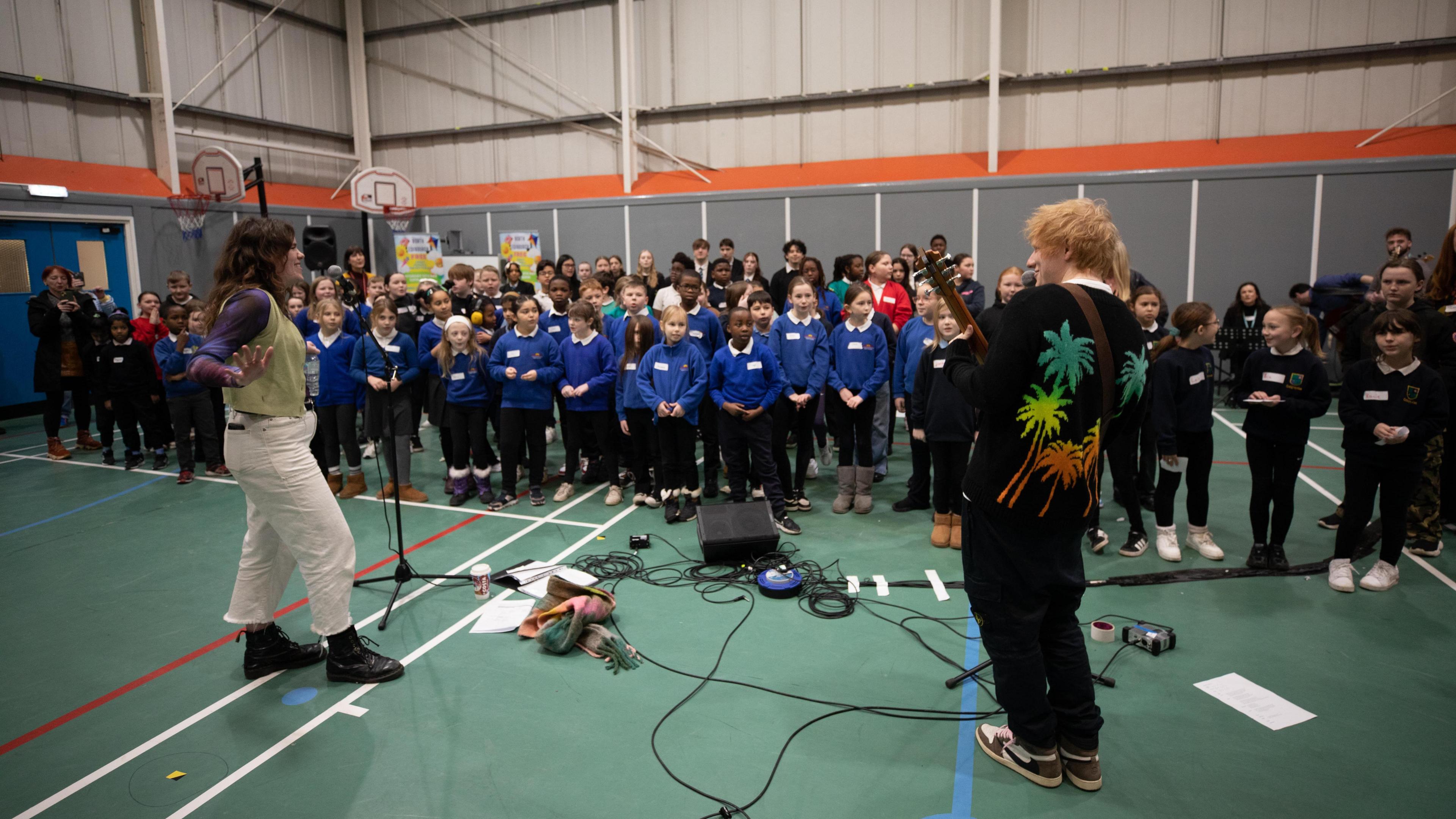 A school gymnasium with green floors and white, red and blue sports markings. Ed Sheeran is standing in the foreground with his back to the camera. He has orange hair and is wearing a black top with multi-coloured palm trees on the back. Another adult is wearing a green vest with white trousers and dark-coloured boots. In the background is a choir of children. Most in the front row are wearing blue jumpers and dark trousers.