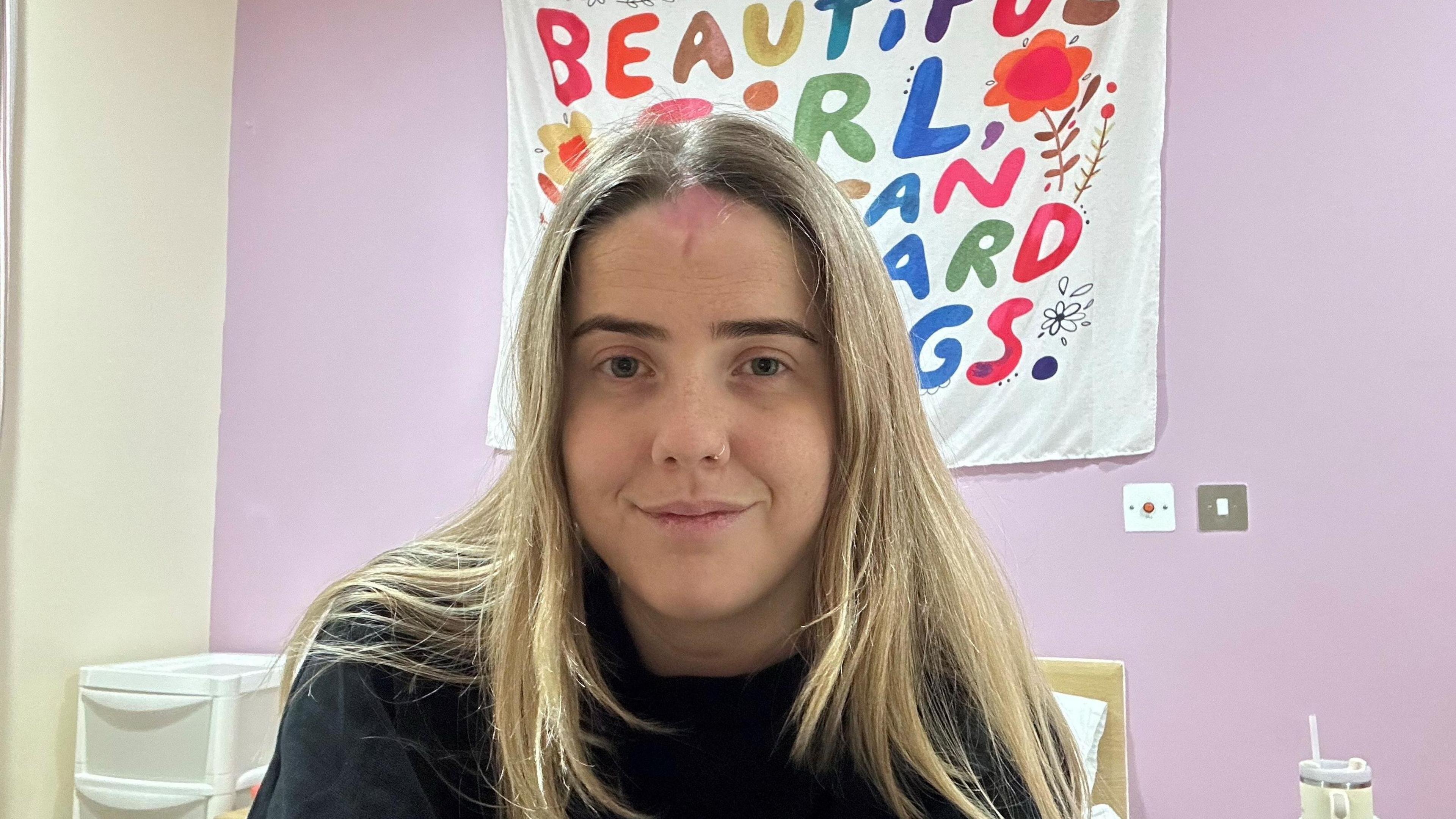 Rachel, who has blonde hair and is wearing a black jumper, sits on a bed with a set of portable draws beside it. A white banner on a purple wall behind her is partly obscured by her head, but the colourful letters on it appear to include the words 'Beautiful girl'