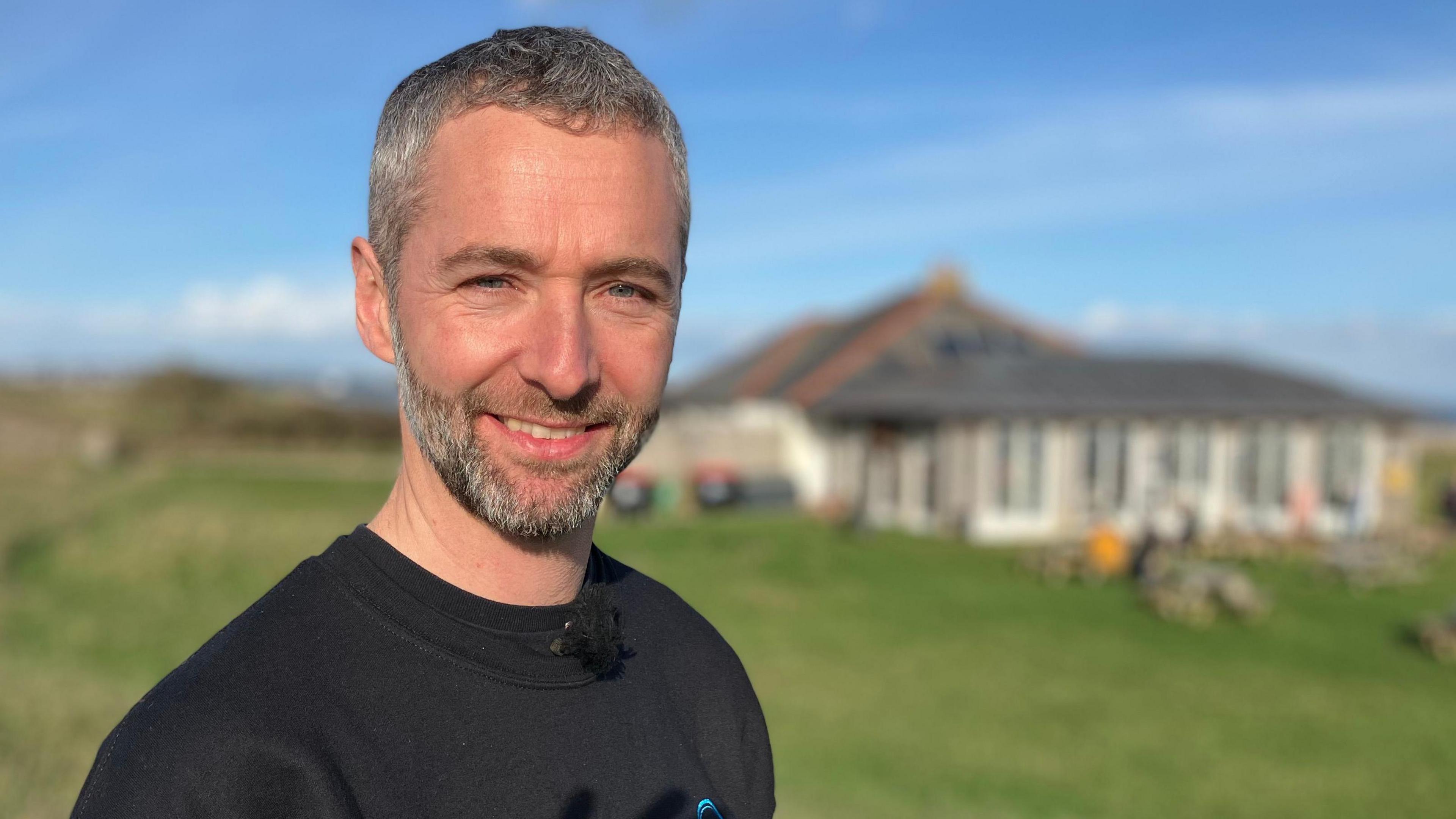 A man stands in front of a cafe building on the coast in Devon. It is sunny, with people sitting at tables. The man wears a black top and is smiling.  