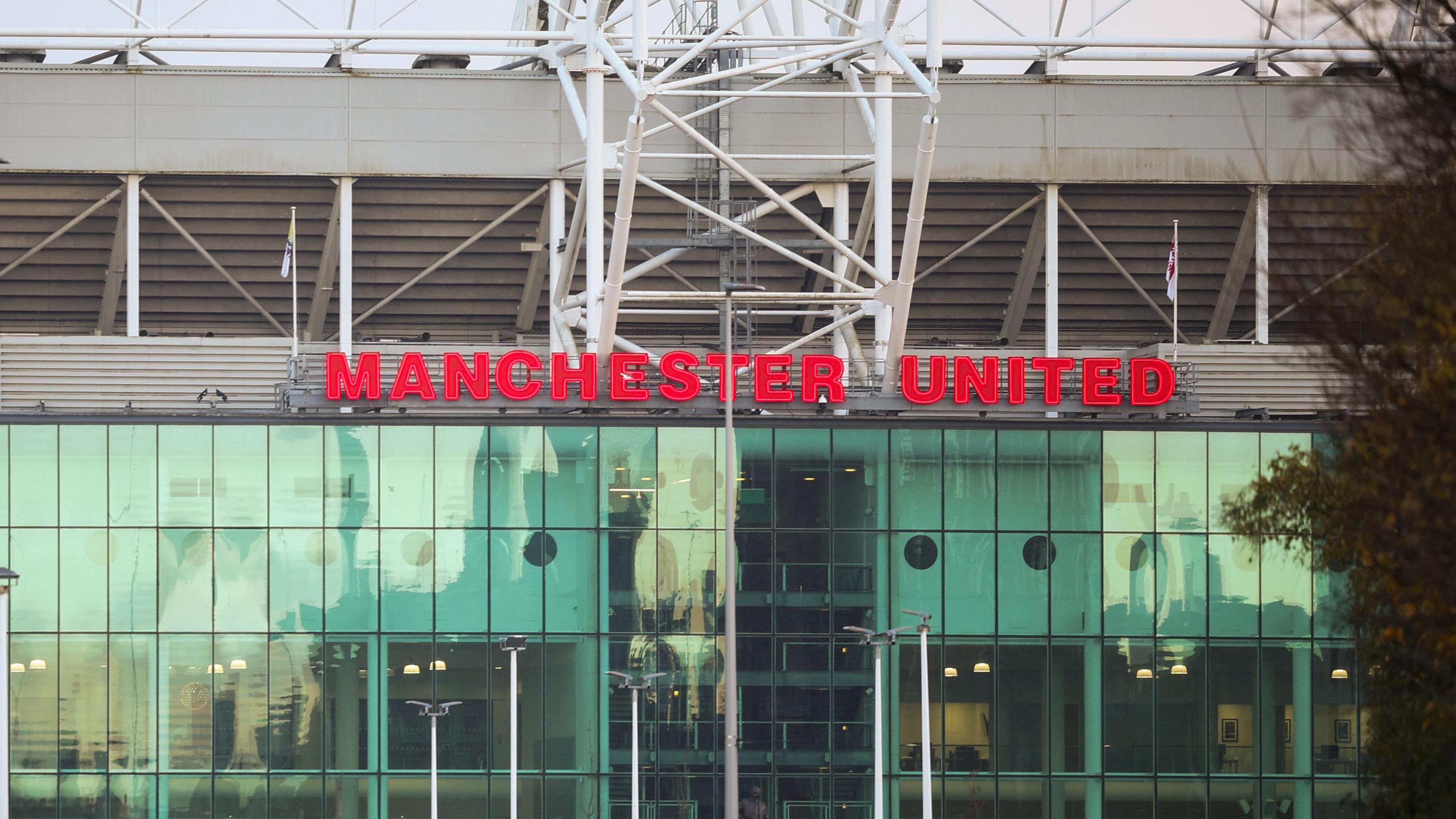 A woman in a blue rain coat with a black rucksack crosses a road, with the Old Trafford stadium seen towering behind her. 