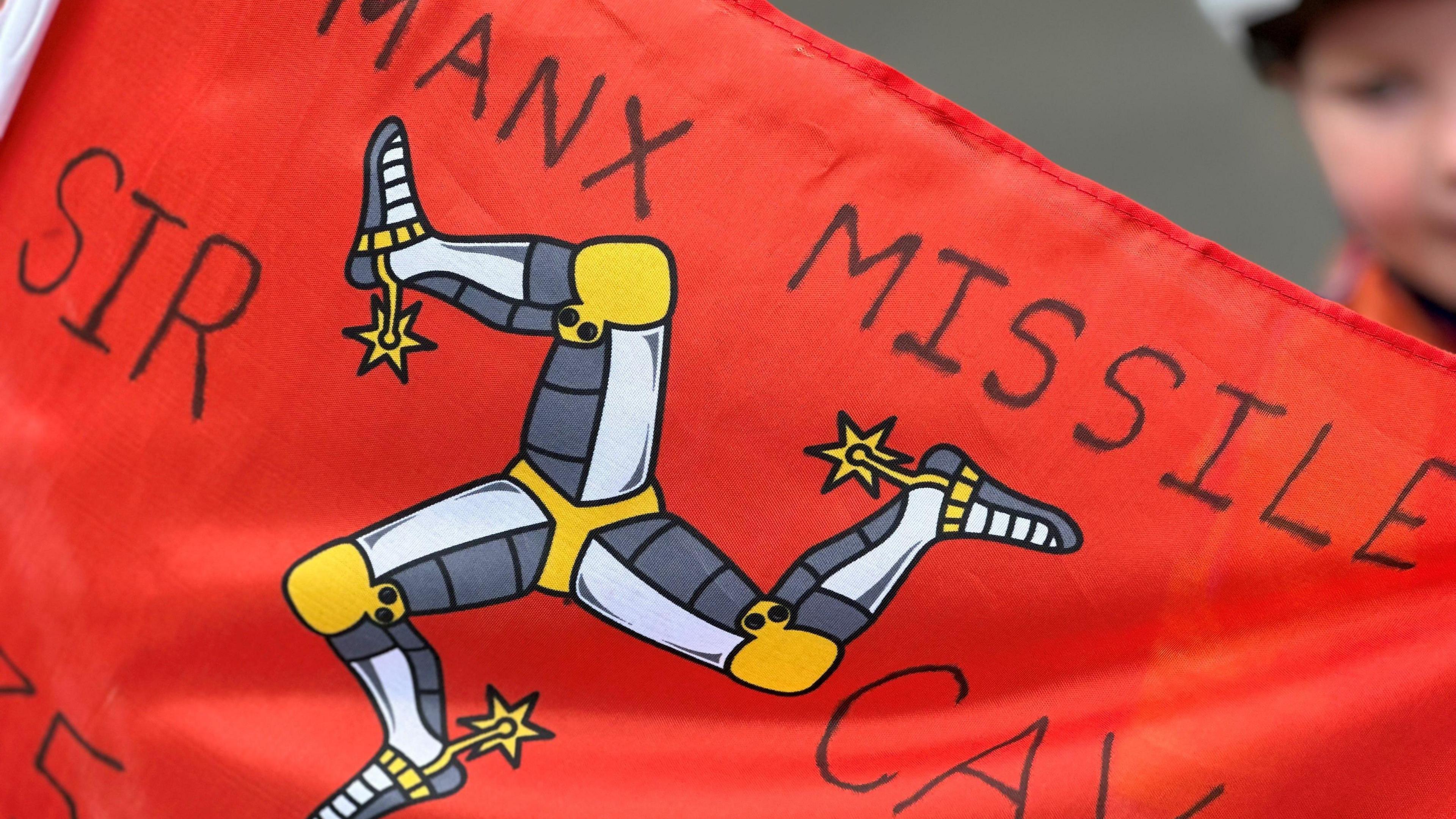 A red Manx flag featuring the three-legs-of-Mann, with the word Manx Missile Sir Cav written on it.