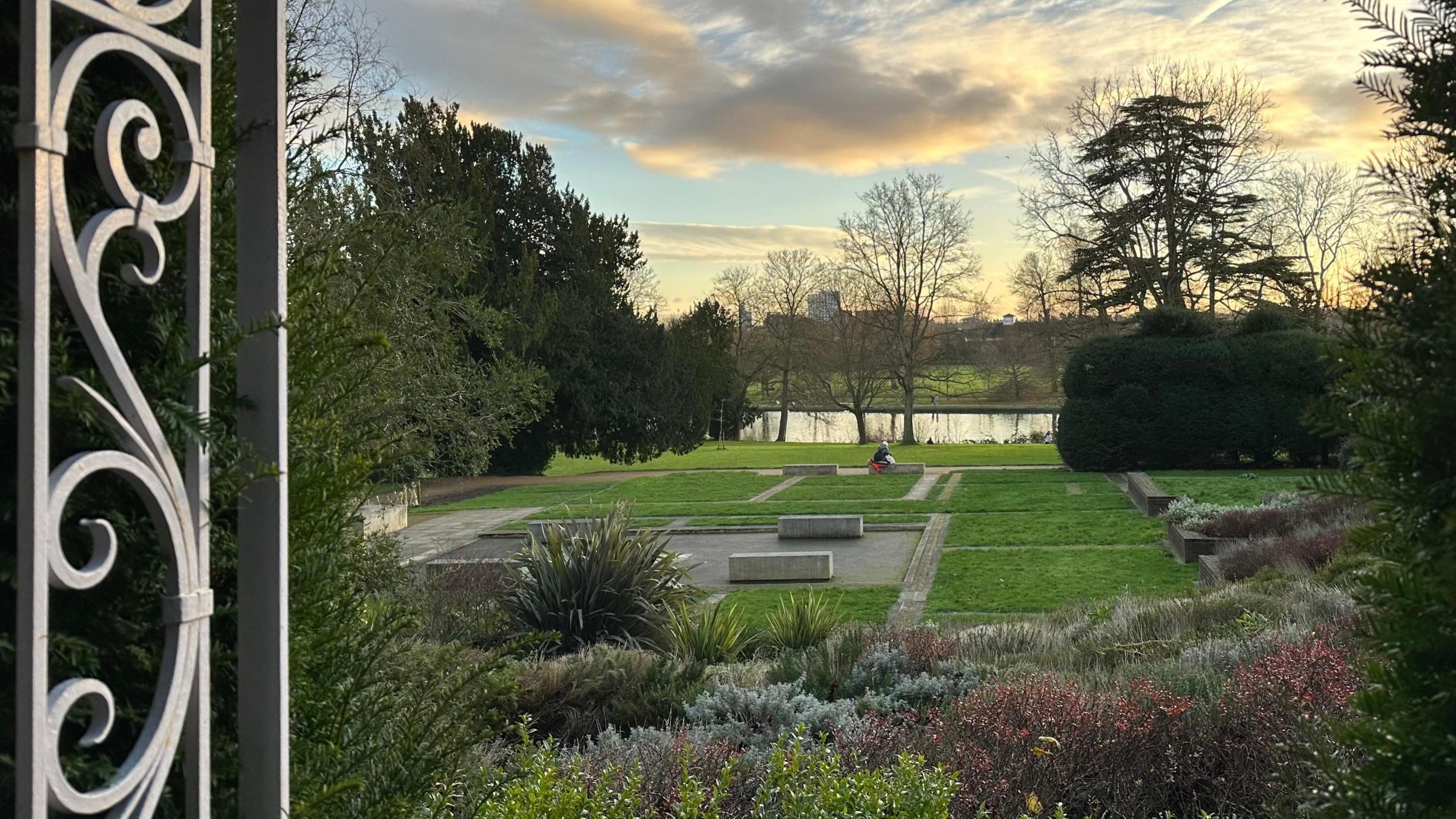 A garden lies in the centre of the picture with a vibrant green lawn laid out in squares. In the foreground, there are several shrubs and bushes with an ornate gate open on one side. In the background, you can make out the river with trees lining both banks.