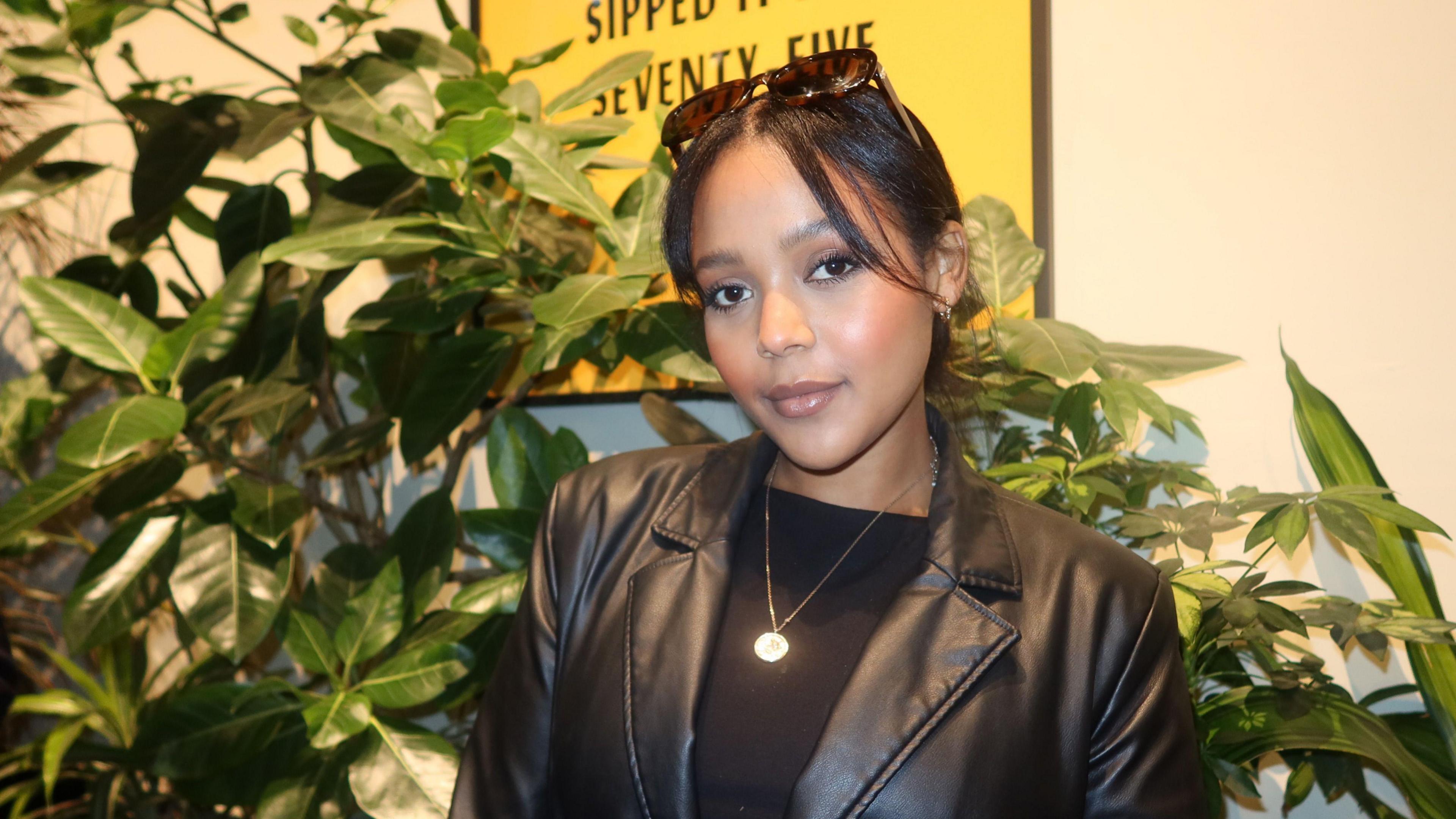 Winta Zesu wearing a black leather jacket stands in front of plant.