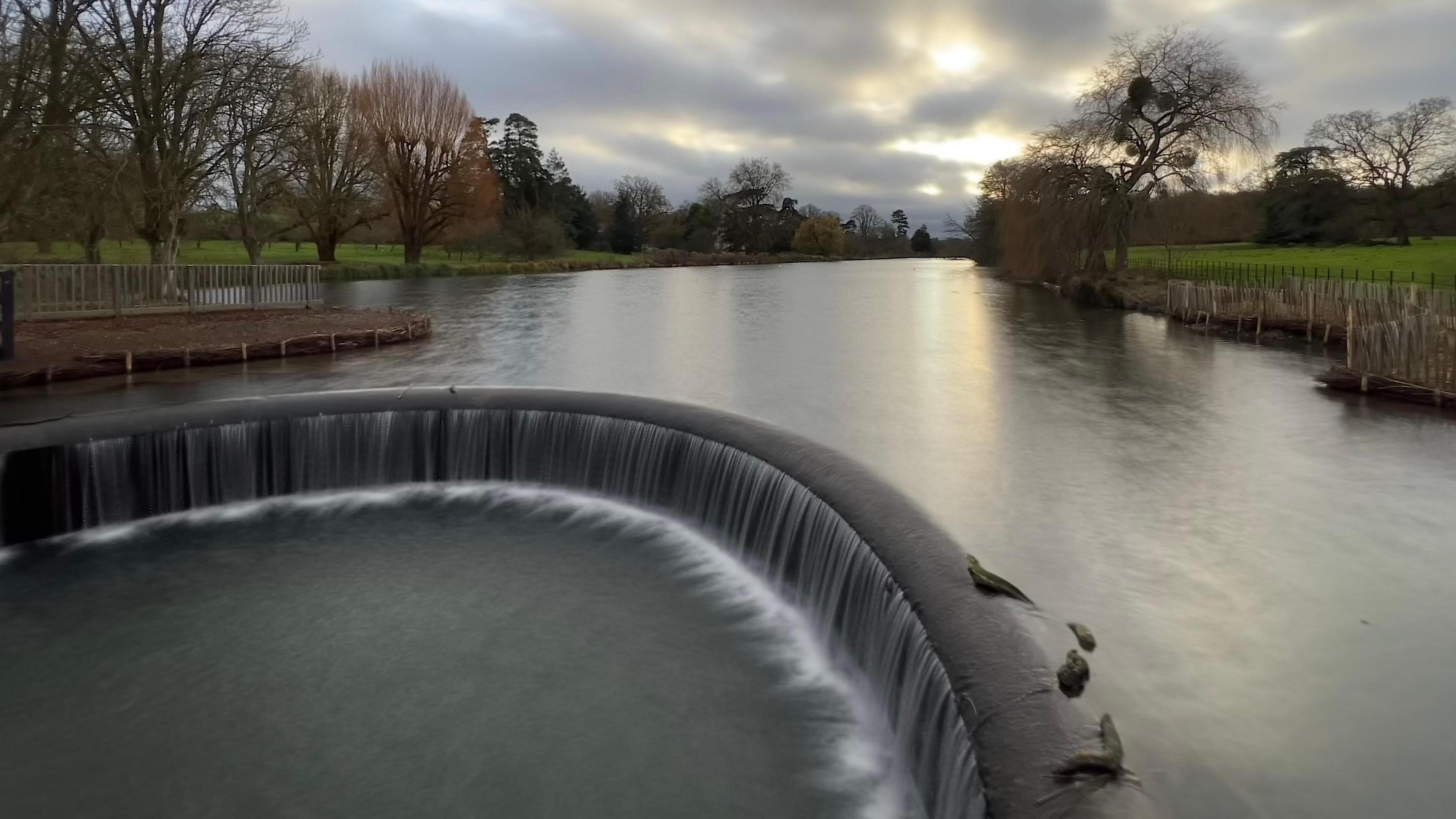 A weir stands in the middle of a river with water flowing over its edge and dropping down into a separate pool of water. The banks on either side of the river are lined with trees.