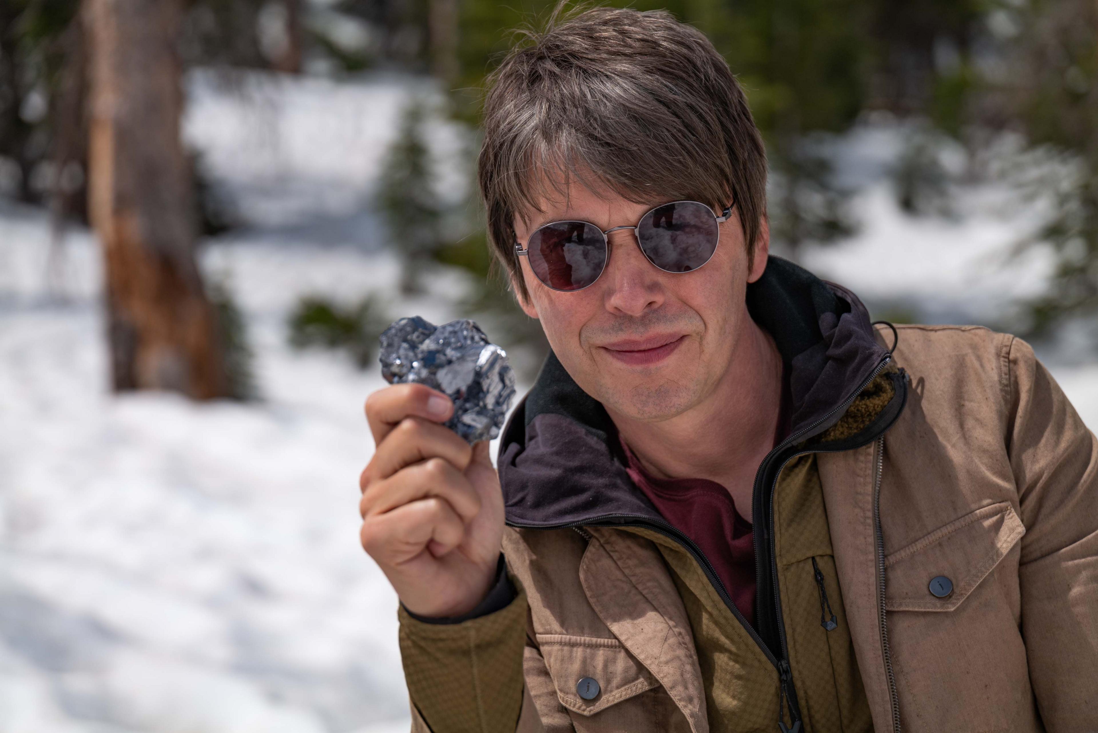 Prof Brian Cox stares straight ahead. In this close-up image, he is wearing a brown jacket and has sunglasses on. He holds a silver rock in his hand made out of lead sulphide. 