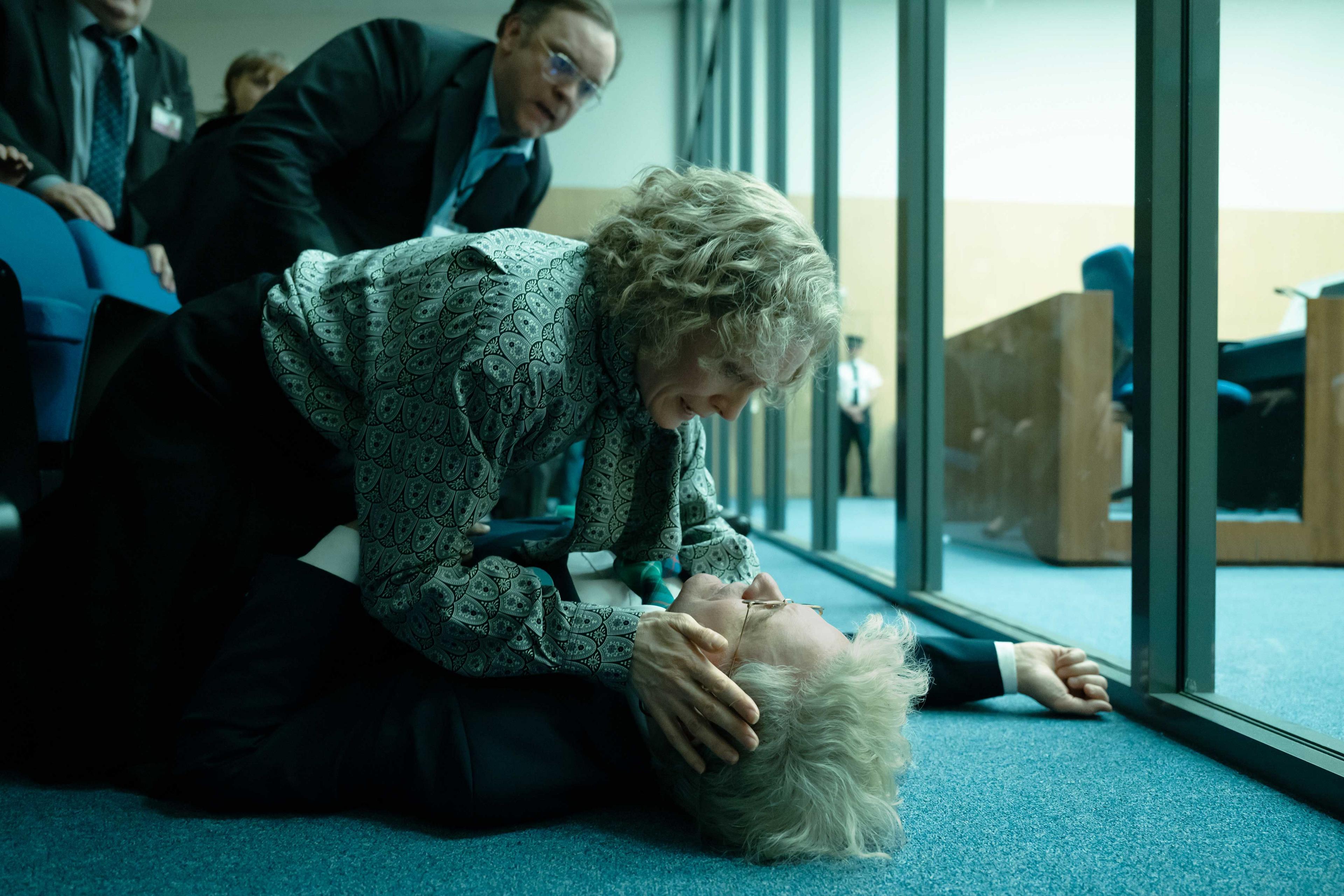 Colin Firth lies on the ground next to a glass partition while a woman holds his head and another man leans over looking concerned