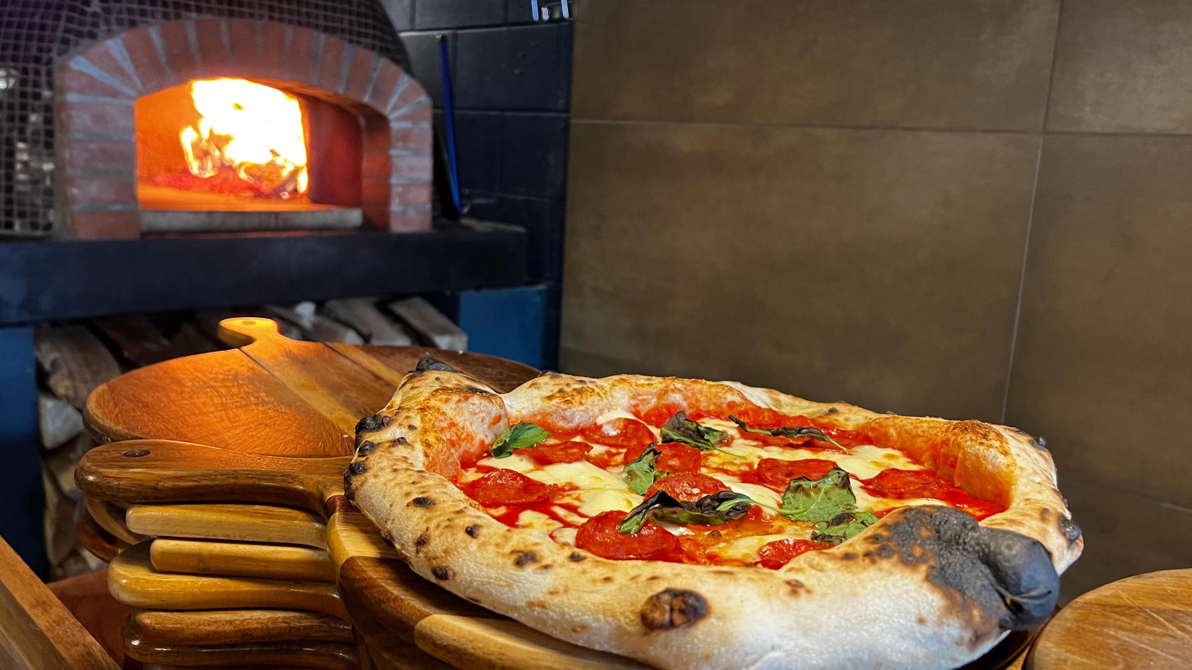  A fresh pizza with salami and basil sits on wooden boards, just in front of a wood-fired oven, with flames licking the roof