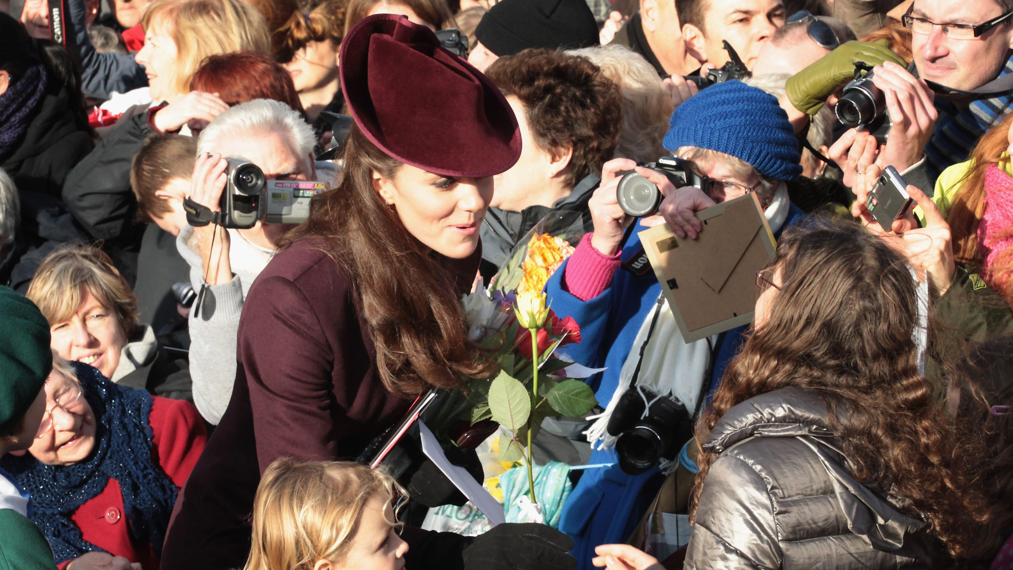 A woman with a burgundy hat and coat is chatting to people in a crowd. She is being given flowers. People all around her are taking photos of her with cameras and video cameras. The crowd is very tightly packed around her with people of all ages. The woman is smiling and engaged in conversation.