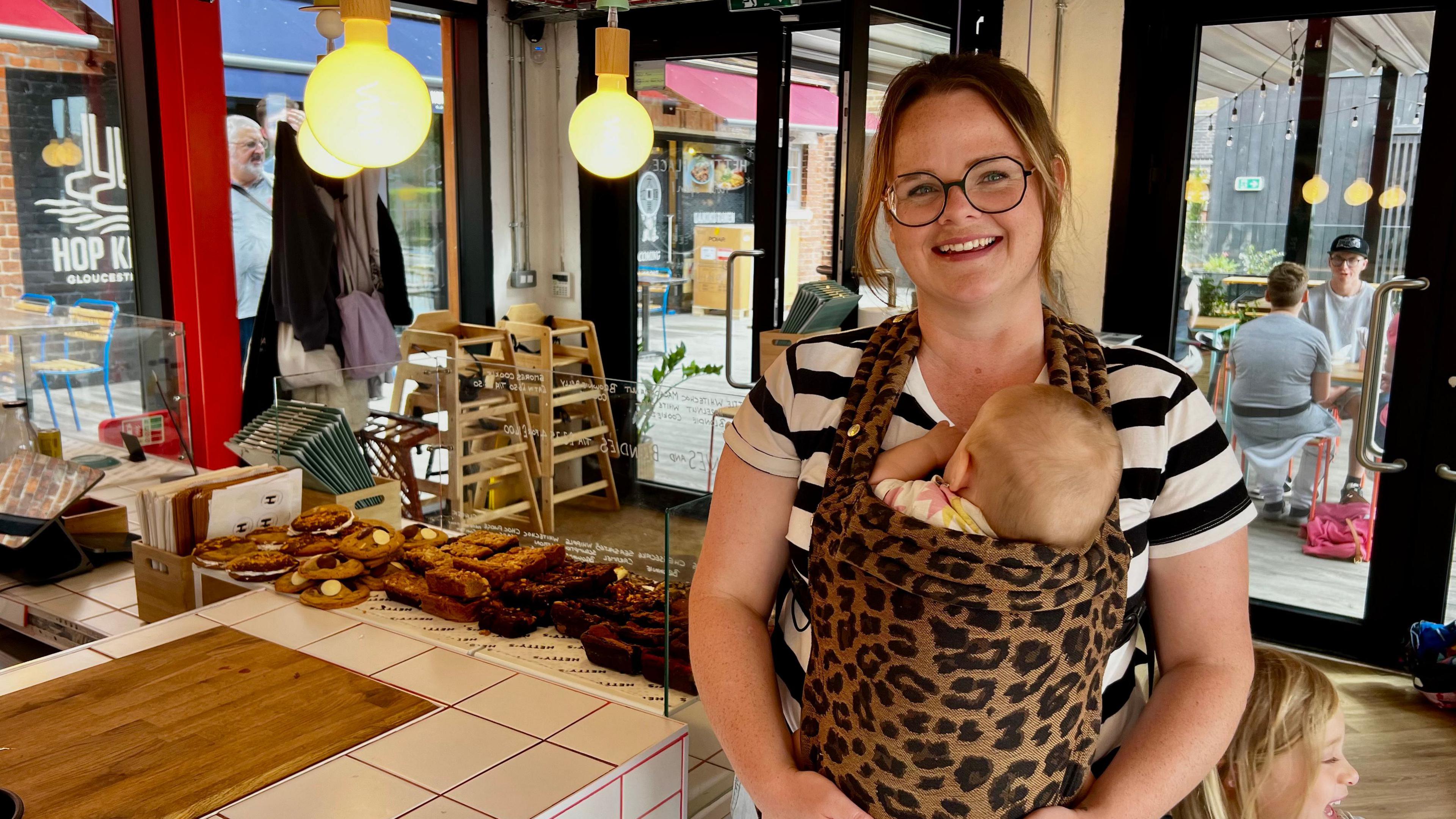 Hetty Kitt stands in front of a display of brownies, wearing a striped blue and white T shirt. Her baby daughter nestles in a sling on her chest