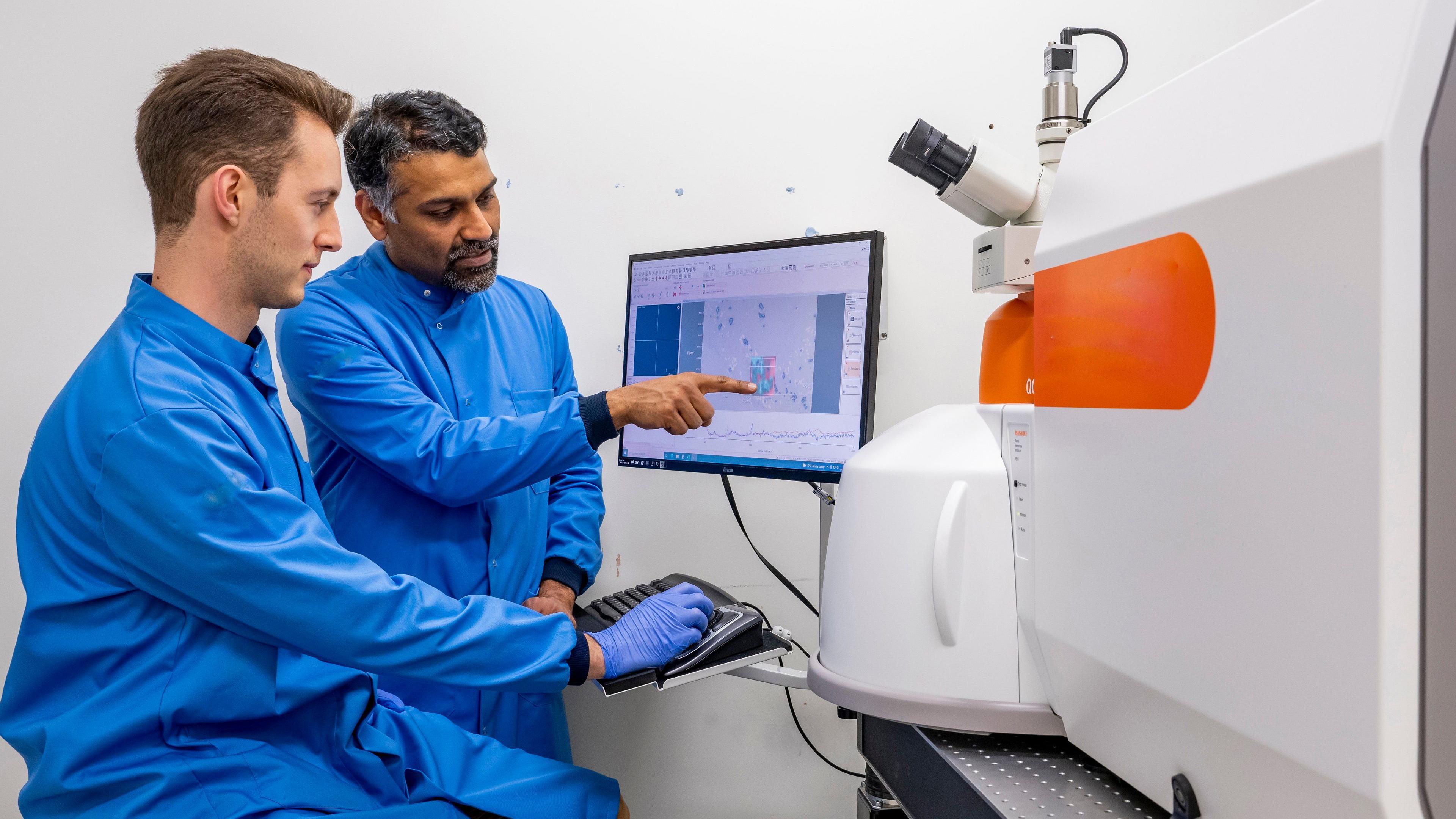 Prof Sumeet Mahajan wearing a blue medical gown alongside another man in a medical gown. Mr Mahajan is pointing at a computer screen with figures on and the other man is using the computer keyboard