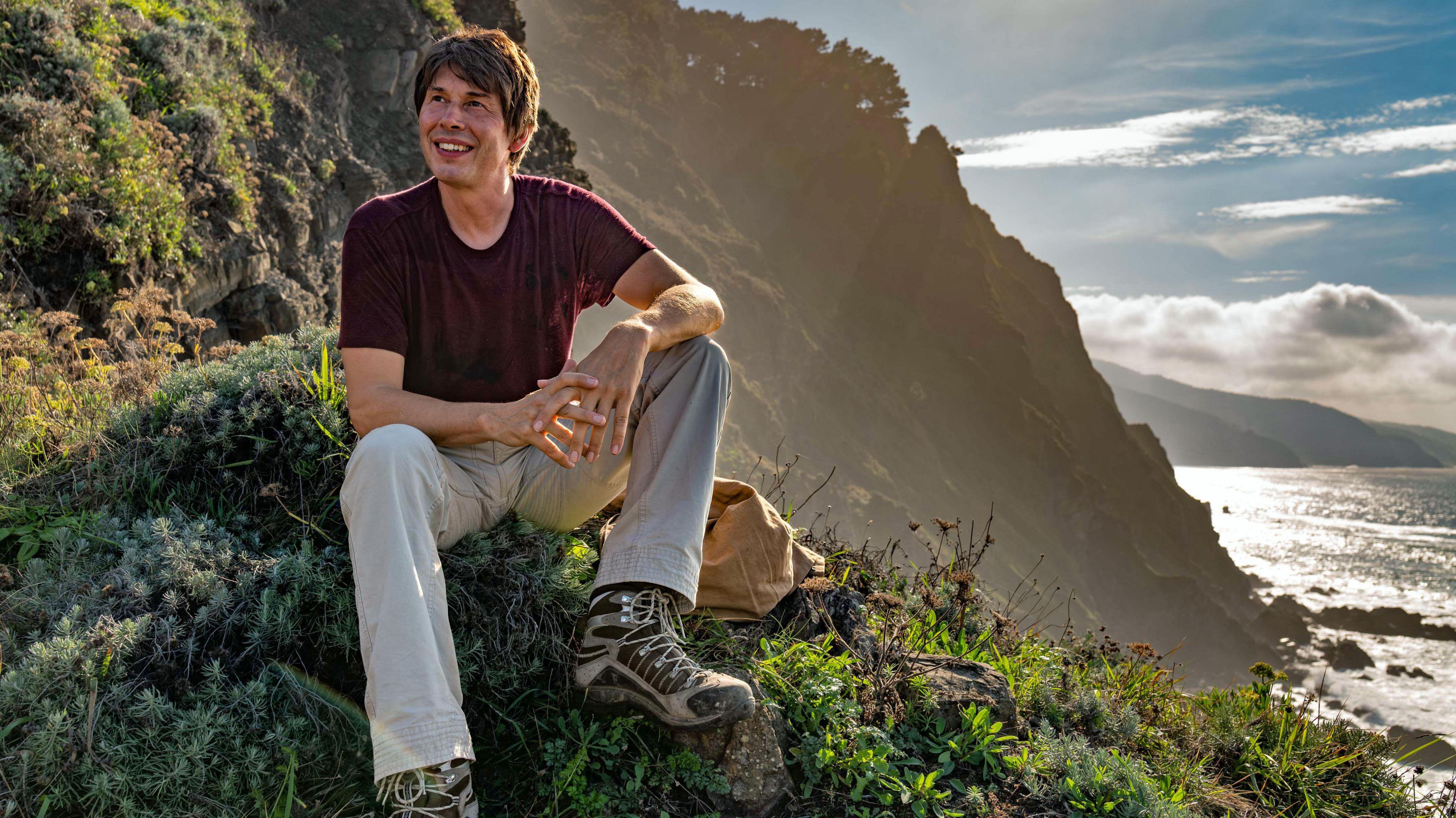 Prof Brian Cox sits on a tree-covered hillside, with the sea on the right hand side of the picture. He is wearing a burgundy t-shirt, grey trousers and walking boots. He is smiling while looking into the distance. The Sun is shining brightly in the background.