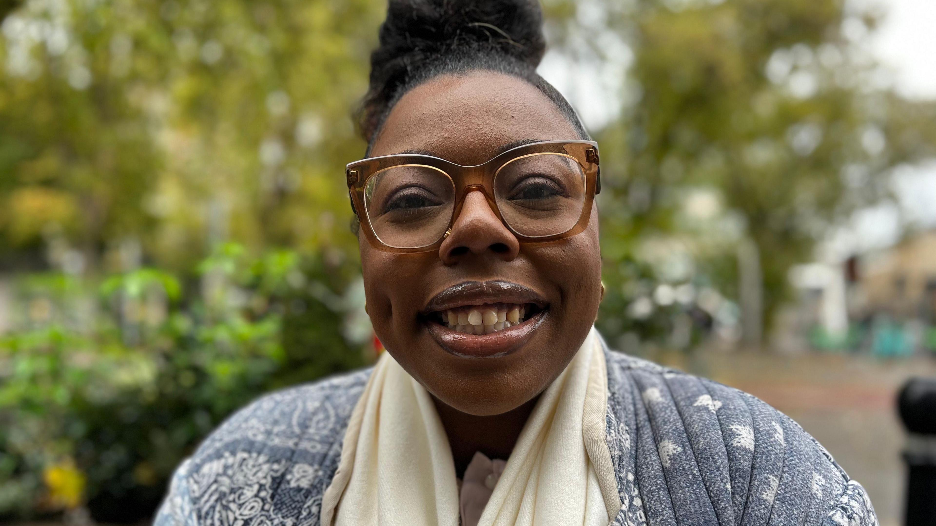 Amara Lawrence is looking at the camera with a wide smile on her face. She is wearing a blue jacket and a white scarf. Behind her are trees out of focus.