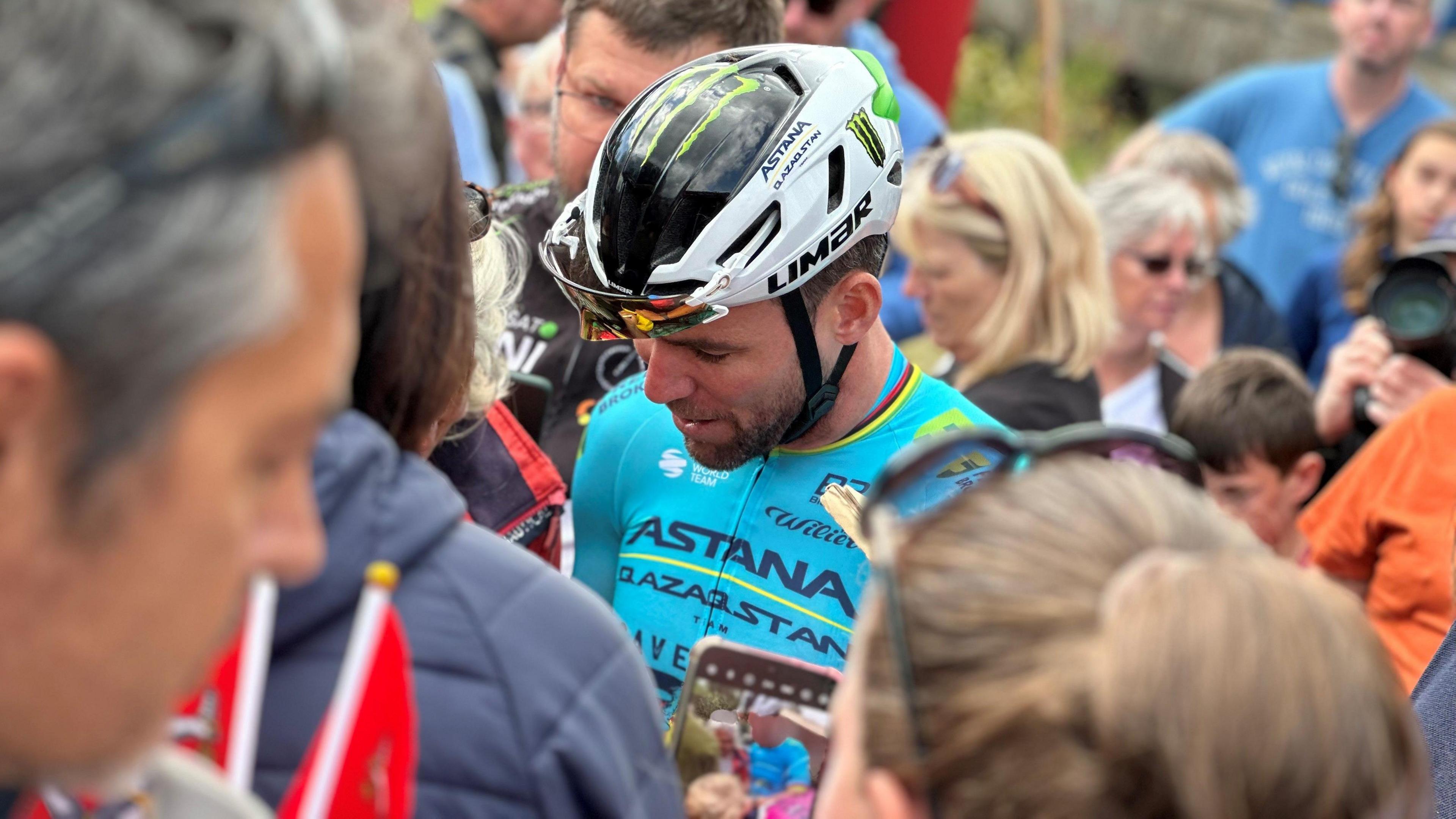 Mark Cavendish in wearing light blue cycling top and white helmet in the middle to a crowd of people.