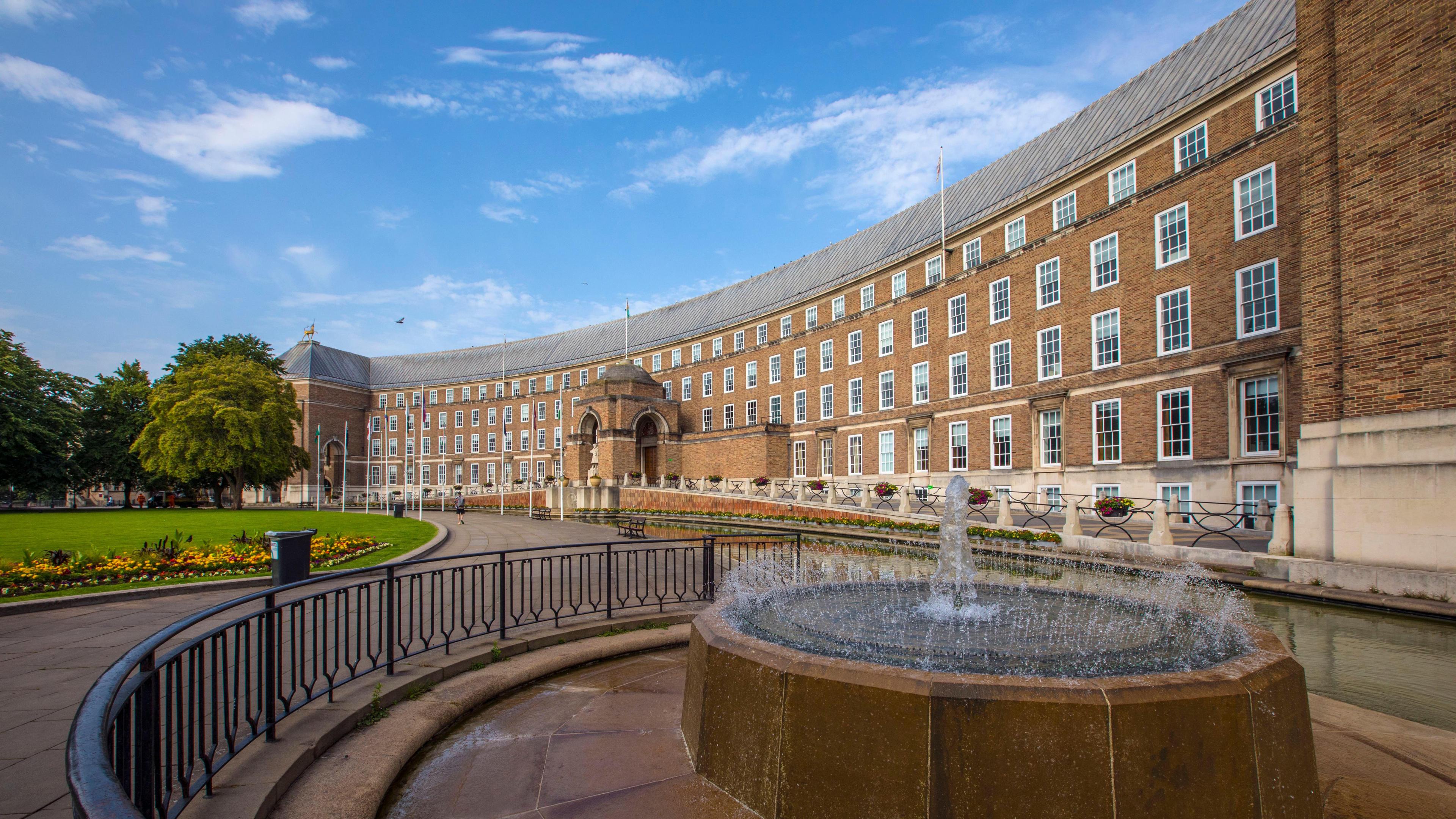 Bristol City Council City Hall. It is a wide, brown brick building with lots of windows. Flag poles are along the front of the building and a fountain can also be seen. 