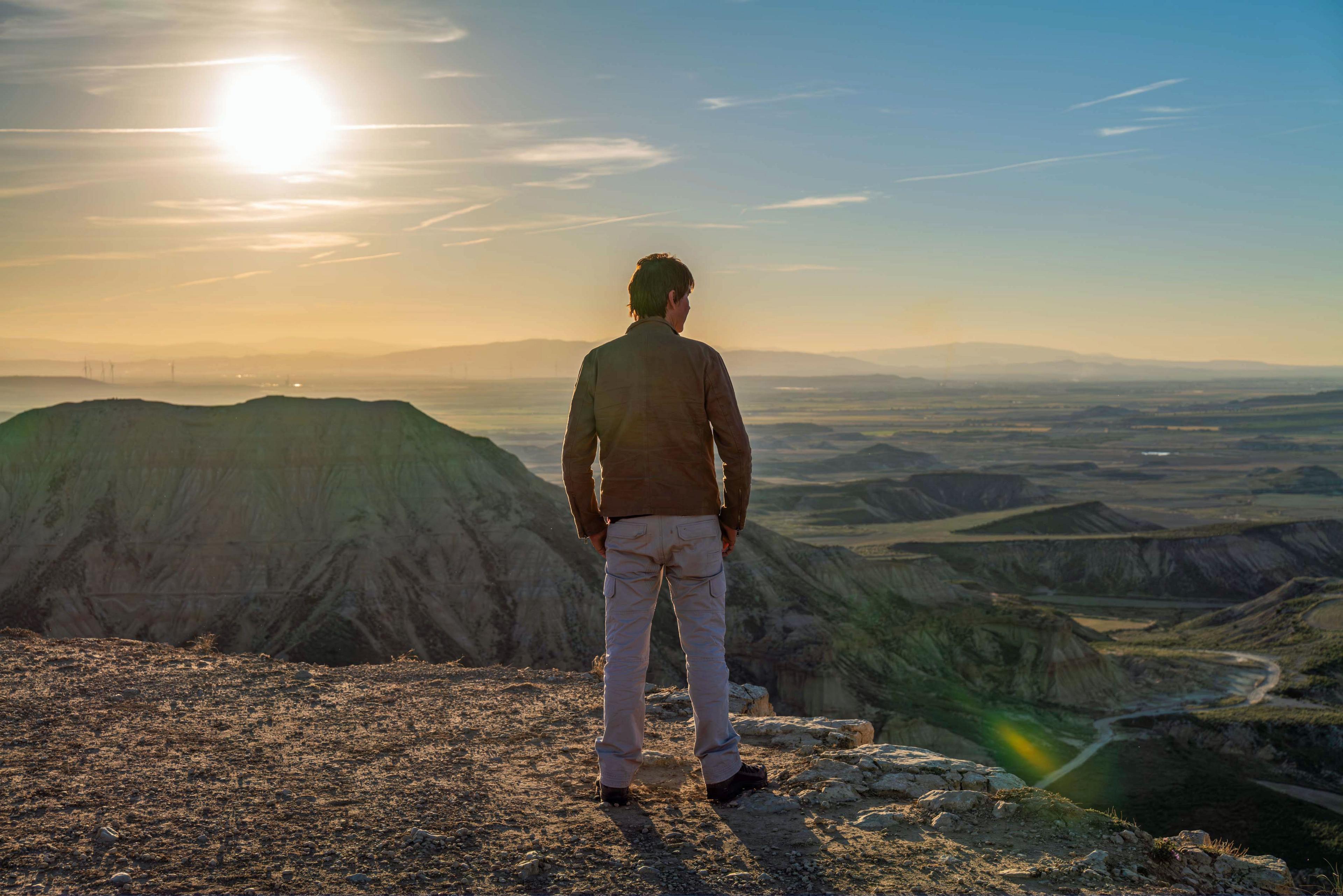 Prof Brian Cox is pictured from behind, looking out on to the horizon from the top of a mountain. He is wearing a brown jacket and grey trousers. Green hills can be seen in the distance and the Sun is shining brightly.