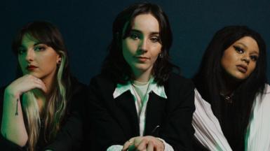 Cherym, made up of L-R Allanagh Doherty, Emer McLaughlin and Hannah Richardson. The band are dressed in monochrome and sit posing on a black sofa against a dark blue backdrop. 