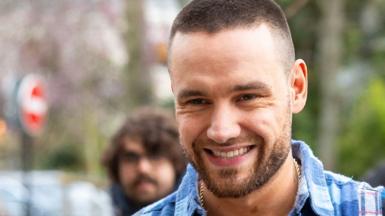 Liam Payne smiles as he looks at the camera while outside and wearing a checkered light blue shirt, the image is cropped close as a headshot, taken in Paris in March