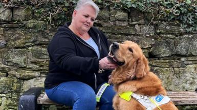 Woman with short grey hair sits on a park bench in front of a stone wall. She is wearing a black hoodie and jeans and she is holding a golden retriever's head in her hands lovingly - the dog is wearing a guide dog collar.