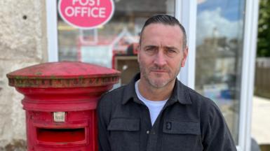Will Mellor stood outside a Post Office leaning on a post box