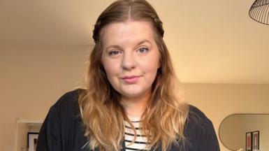 Katie Smith stands in her home, wearing a black shirt over a striped t-shirt. She has shoulder length dark blonde hair and a nose ring. 