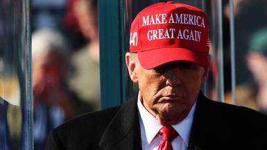 Donald Trump looks down as he walks towards camera while wearing a 'Make America great again' red cap, and a suit with a red tie, at a campaign event