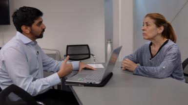 A man and a woman sit across a grey table from each other. The man, on the left, has a laptop open in front of him and is talking while gesturing with his right hand. The woman, on the right, is looking at him, listening.