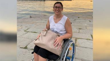 A woman in her twenties sits in a wheelchair with a large bag on her lap next to water, she smiles and looks towards the camera