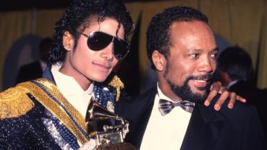 Michael Jackson and Quincy Jones are pictured together at the 1994 Grammy awards, Jackson is wearing a blue and gold sparkley suit with sunglasses on and holding a grammy award, and Jones looks away from camera wearing a suit and bow tie.