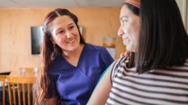 Two women smiling at each other. One is a medical practitioner and is wearing blue scrubs, the other is a patient who seems to be being examined.