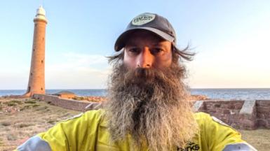 Sandy Duthie, man with large greying beard, with a lighthouse in the background, and sea, and blue skies.