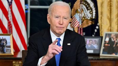 Joe Biden sits at a desk with photos and a US flag behind him and points towards the camera.