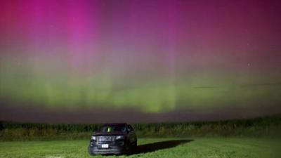 Pink and green Northern Lights hang in the sky above a car in a field