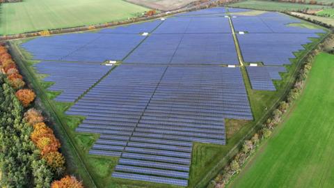 A drone picture shows a huge solar farm near the village of Great Wilbraham, near Cambridge, from above. Thousands of panels cover a huge field. The solar farm is surrounded by fields and trees.