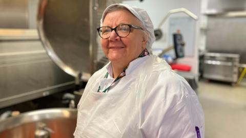 Celia wearing white chef's overalls and glasses smiling at the camera