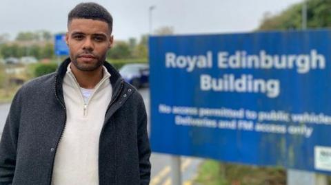 Christopher MacRae, wearing a cream fleece and grey coat, standing in front of a sign for the Royal Edinburgh Hospital