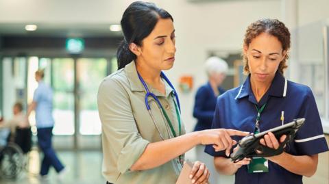 Two female medical staff stand together, one in a blue uniform is holding an electronic tablet and the other woman, wearing a grey shirt and a stethoscope around her neck is pointing at something on the tablet's screen