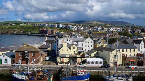 A range of properties in the seaside town of Peel. Boats moored in the inner harbour can be seen in the foreground, with buildings behind them stretch out into the distance. 