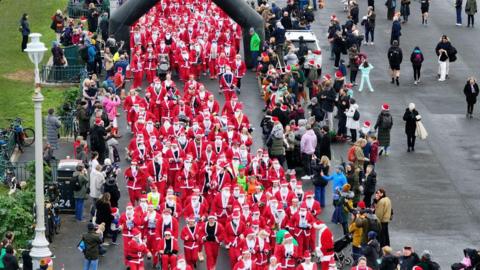 Hundreds of people running on Hove seafront, next to crowds of people cheering them on. All are wearing red and white outfits, a white beard and a Christmas hat.