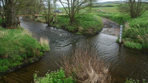 A rural road crosses a river via a deep ford. There is a footbridge to the left, bushes and vegetation, and a white measuring post.