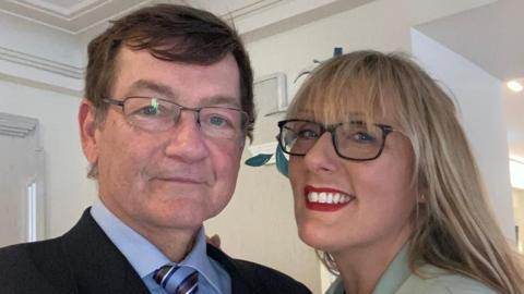 A selfie-style photo of Phil and Juli looking into the camera. Phil is wearing a dark blazer with a blue shirt and stripy tie and Juli is wearing a green coloured top and a green fascinator.