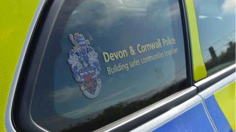 A picture of the Devon and Cornwall police logo on the side window of a police car. The car is bright yellow and blue.