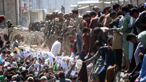 A large crowd of people waving papers in front of a line of men with guns.