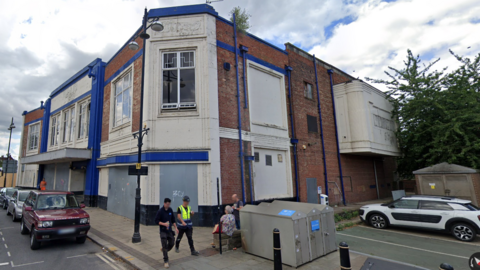 A run-down former cinema building in a town centre, with cars parked alongside.