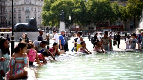 People by a fountain in Trafalgar Square during summer in London. Some are dipping their hands into the water.