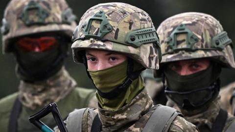 Close headshot portrait of three Ukrainian soldiers, wearing helmets and camouflage, training with the British Army in the UK in August 2024