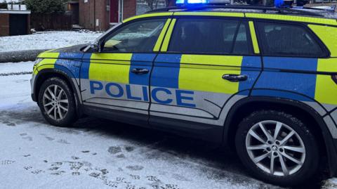 A police car parked at the scene on the shooting. The pictured in cropped in on the car, so no house can be seen. There is snow on the ground.