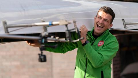 Former rugby player Ed Jackson wears a green sport Relief jacket and helps carry a rowing canoe.