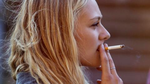 A young woman with long blonde hair smokes a cigarette outside.