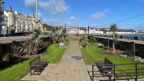 One of the sunken gardens, which is a green space with a large path and benches on either side. The promenade walkway and Douglas Bay are on the right and the Gaiety Theatre and Villa Marina are across the road to the left of the image in the distance.