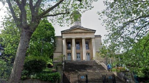 A view of the hall from the street. It is a cream building with several pillars. Leading up to are the building are stone steps and in front of these are black gates. Trees are in the front of the picture and there are also bushes and foliage. 